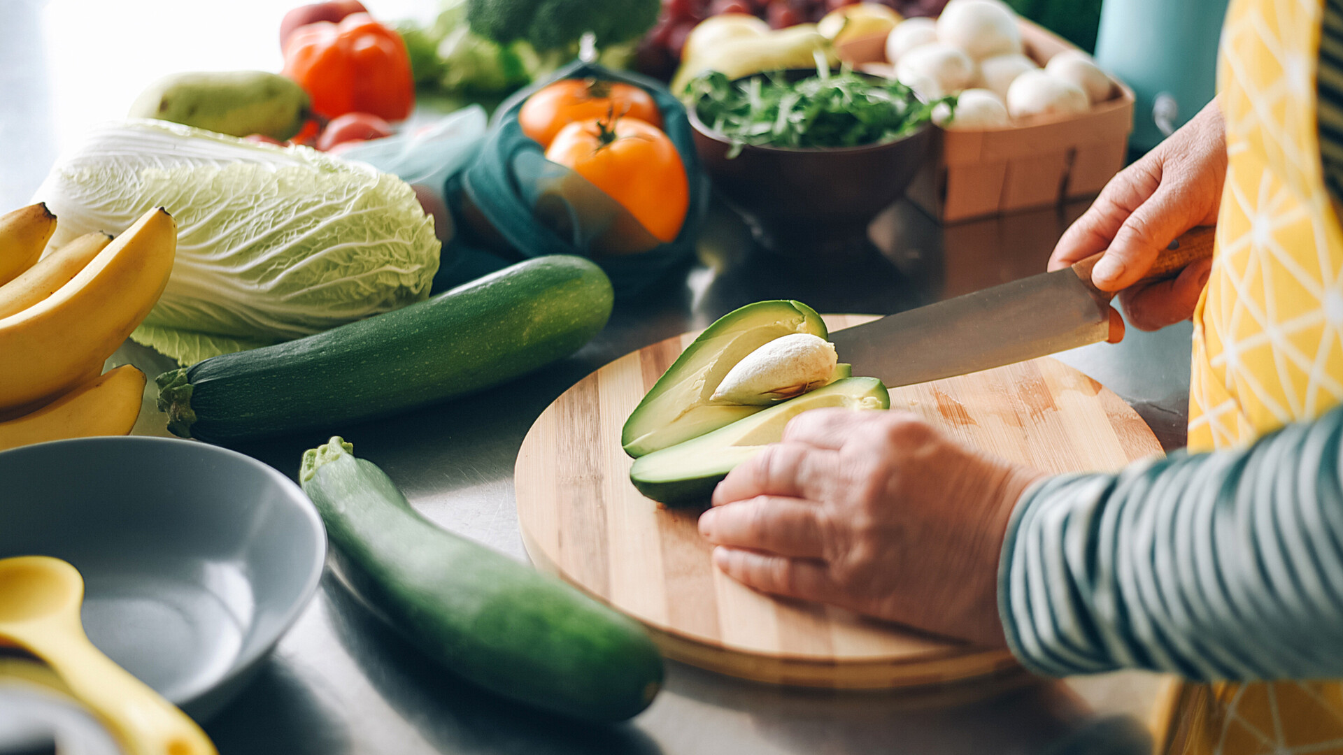 Eine Frau schneidet eine Avocado auf einem Brett. Auf der Anrichte liegt weiteres Gemüse.