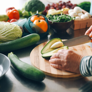 Eine Frau schneidet eine Avocado auf einem Brett. Auf der Anrichte liegt weiteres Gemüse.