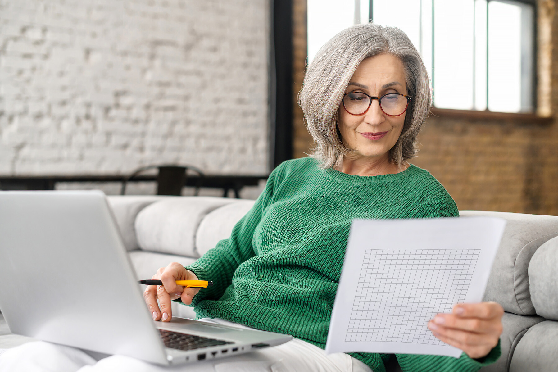 Frau mittleren Alters sitz mit Laptop auf der Couch und macht den Amsler-Gitter-Test