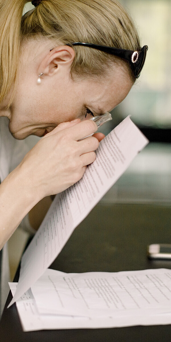 Frau mittleren Alters sitzt am Schreibtisch und liest mit Lupe ein Dokument